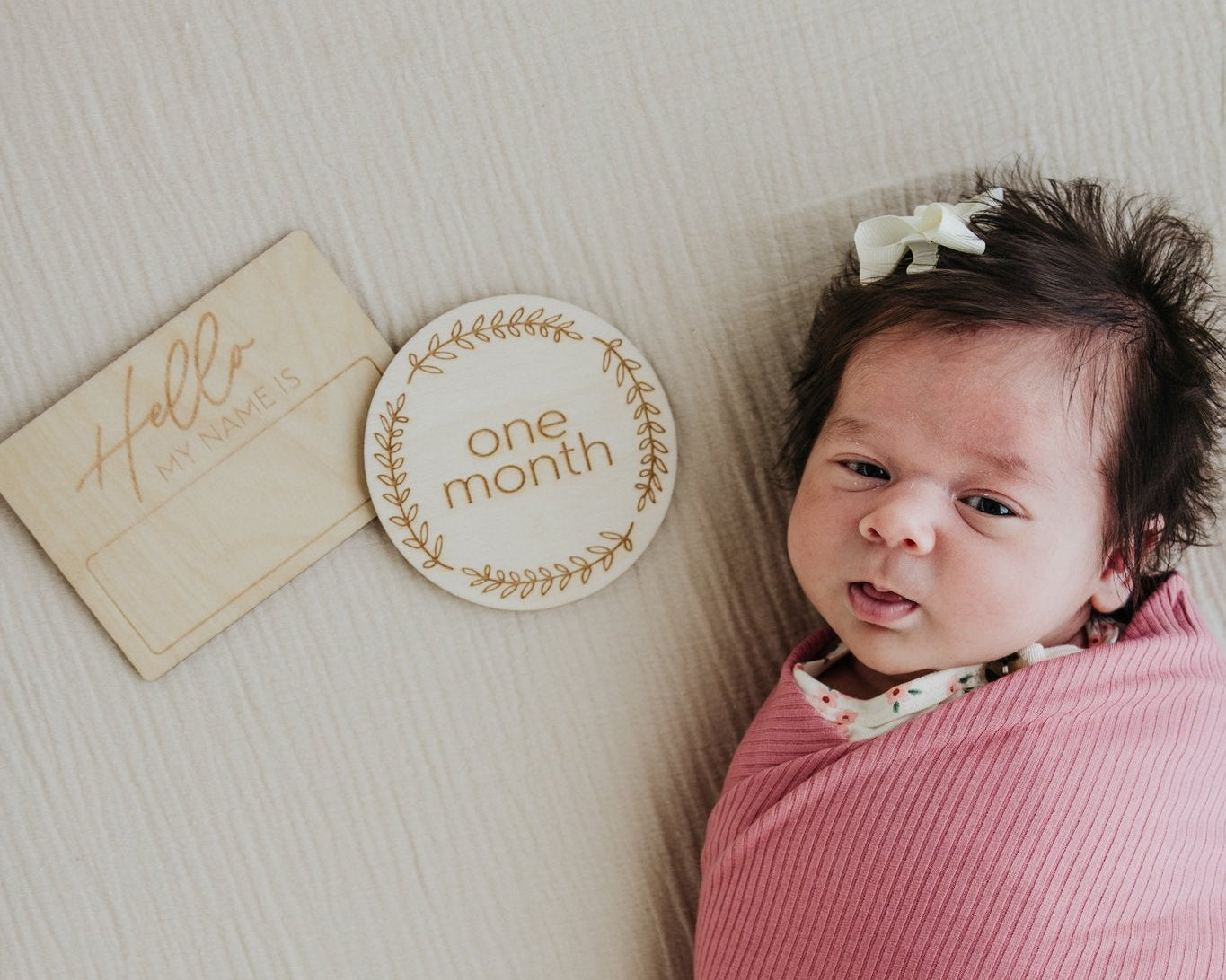 Newborn baby wrapped in pink swaddle with 'one month' milestone plaque on a white surface